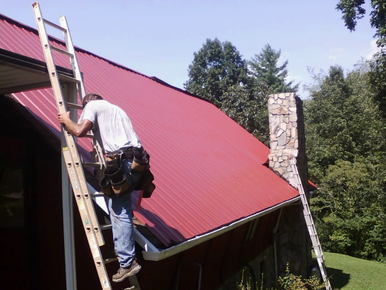 Roofer on a ladder beside a red metal roof and stone chimney — Soffit & Fascia Repair by Roofing Brothers in Gardena
